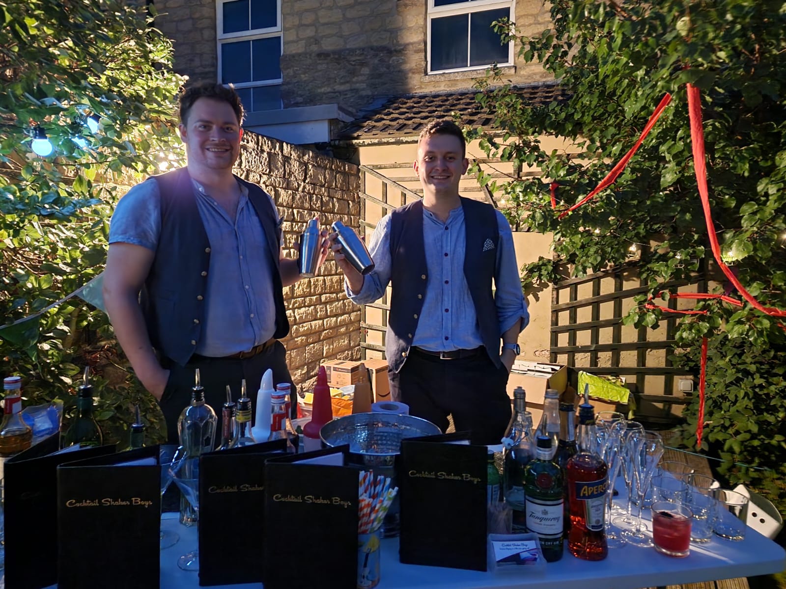 Duncan and Tom Shaking This is an image of our head Mixologist, Duncan, and another mixologist, Tom, holding cocktail shaker, at one of our clients' parties. The party is outside, in a garden, at night time. There is a table in the foreground with glassware, spirits, and menus on it.
